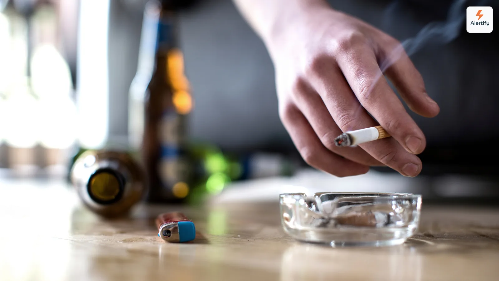 Huésped sosteniendo un cigarrillo encendido sobre un cenicero de cristal junto a botellas vacías en una mesa