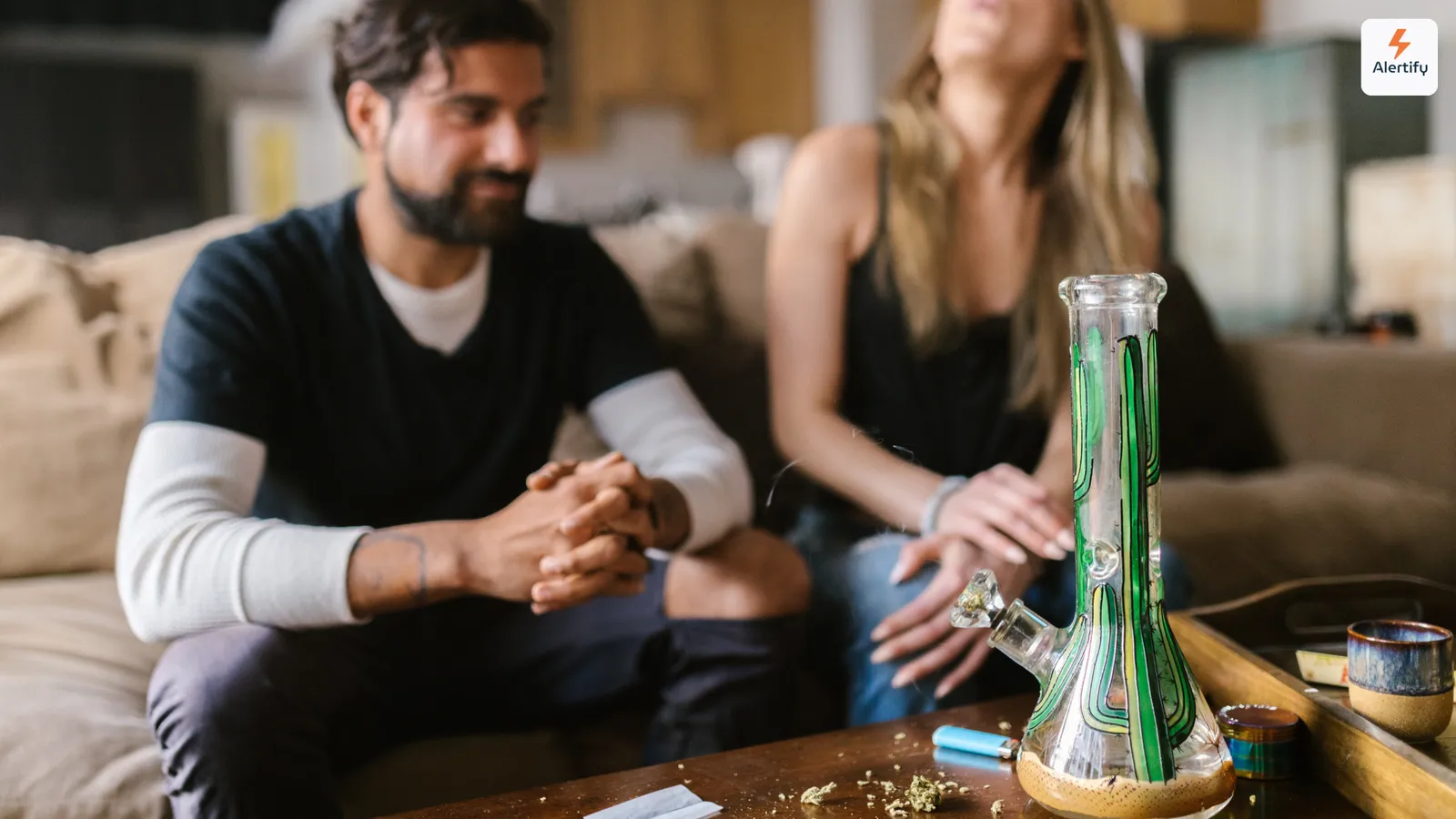 Dos huéspedes sentados en un sofá en el salón de un alquiler, con una pipa de agua de cristal, un mechero y un moledor sobre la mesa de centro frente a ellos.