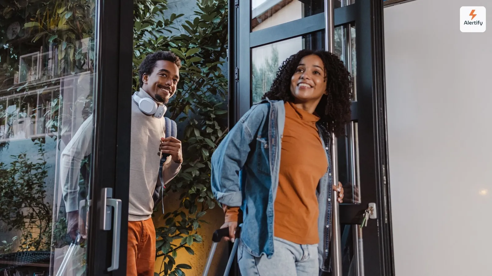 Dos huéspedes llegando a un alquiler con maletas con ruedas, sonriendo mientras cruzan una puerta de cristal.